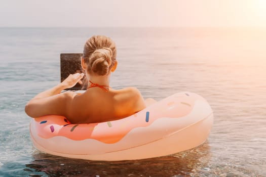 Woman freelancer works on laptop swimming in sea on pink inflatable ring. Pretty lady typing on computer while floating in the sea on inflatable donut at sunset. Freelance, remote work on vacation