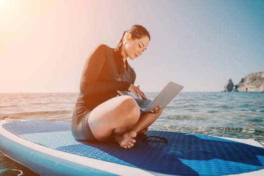 Digital nomad, woman in the hat, a business woman with a laptop sits on the rocks by the sea during sunset, makes a business transaction online from a distance. Freelance, remote work on vacation.