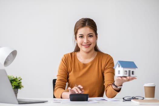 A woman performs calculations related to real estate transactions, holding a model house and using a calculator in a modern office environment.