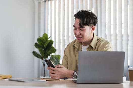 Young male businessman working online with a laptop and smartphone in a modern office environment, showcasing productivity and technology.