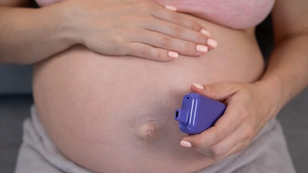 A pregnant woman smokes a vape while sitting on the couch. Close-up of the abdomen