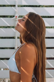 A woman with long brown hair is standing in front of a white wall. She is wearing a white bikini top and is smiling