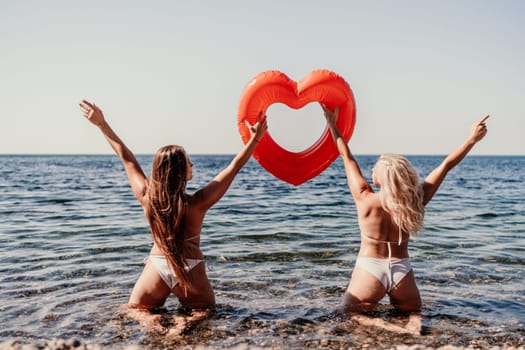 Two women in bikinis are holding a heart-shaped inflatable raft in the ocean. The scene is playful and fun, with the women enjoying their time in the water