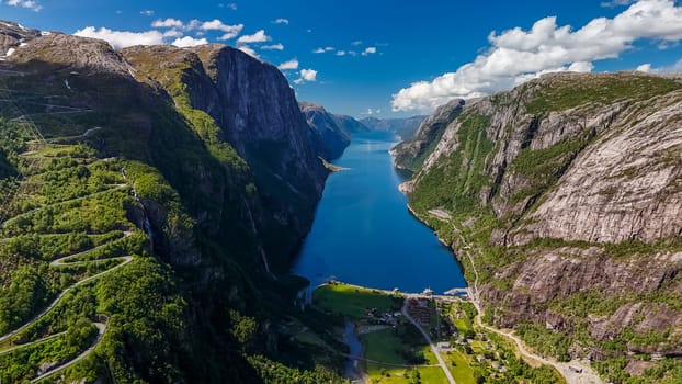 An aerial view of a winding road snaking through the lush green mountains of Norway, with a stunning fjord in the foreground.