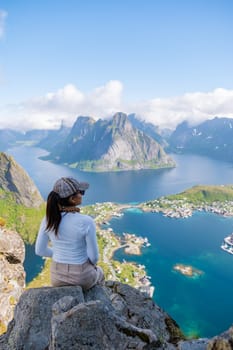 A woman sits on a cliff, overlooking the stunning fjords and mountains of Norway Lofoten Islands. The view is breathtaking, with clear blue water and lush green hills. Reinebringen, Lofoten, Norway