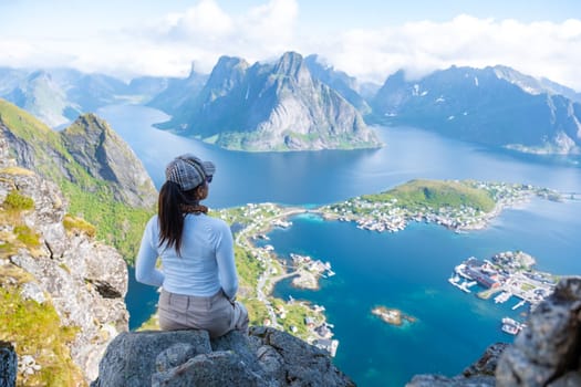 A woman sits on a rocky cliff overlooking a stunning view of the Norwegian fjords, with mountains, clear waters, and a small town below. Reinebringen, Lofoten, Norway