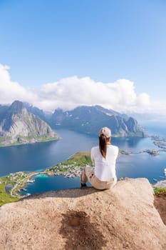 A woman sits on a cliff overlooking a Norwegian fjord, surrounded by stunning mountains and blue waters. beauty and serenity of the Norwegian landscape. Reinebringen, Lofoten, Norway