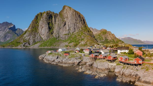 village in Norway with red wooden houses on stilts, nestled against dramatic cliffs, with calm blue waters and a sunny sky. Hamnoy fishing village on Lofoten Islands, Norway with red rorbu houses