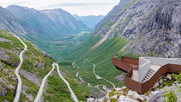 A scenic overlook provides a stunning view of a winding road snaking through the mountains of Norway, with a person enjoying the breathtaking scenery. Trollstigen Road Norway