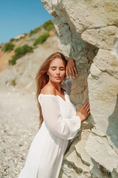 A woman in a white dress is standing on a rocky beach. Concept of serenity and tranquility, as the woman is enjoying the natural beauty of the beach