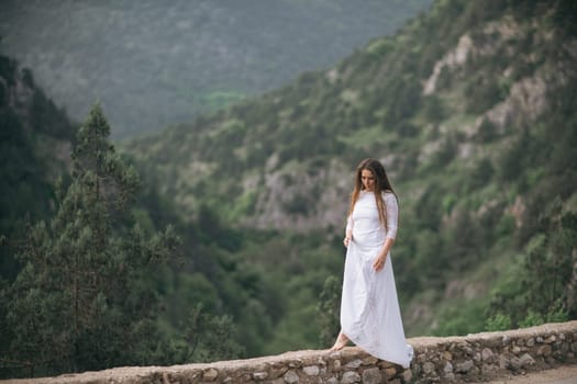 A woman in a white dress is walking on a stone wall in the mountains. The scene is serene and peaceful, with the woman's dress flowing in the wind