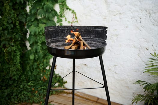 A black round fire pit standing against a green leafy background with burning wood, perfect for outdoor gatherings and cozy evenings.