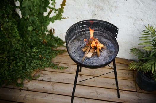 Outdoor fire pit with burning wood on a wooden deck surrounded by lush green plants, creating a cozy atmosphere.