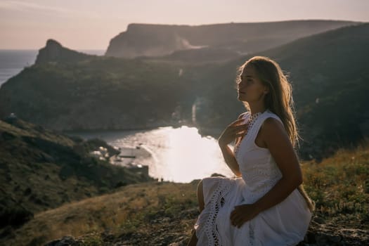 A woman in a white dress is sitting on a rock overlooking a body of water. She is enjoying the view and taking in the scenery