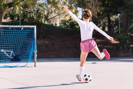 rear view of an unrecognizable young female football player kicking a soccer ball into the goal on an urban field, concept of sport and active lifestyle