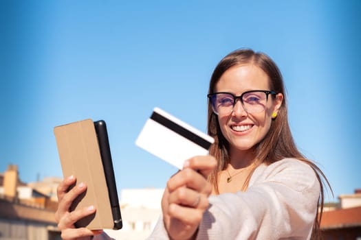 Happy young woman with glasses holding in each hand a credit card and digital tablet. Looking into the camera. Concept: payment in an online store