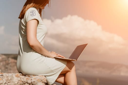 Digital nomad, woman in the hat, a business woman with a laptop sits on the rocks by the sea during sunset, makes a business transaction online from a distance. Freelance, remote work on vacation.