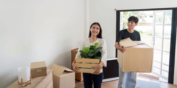A cheerful couple carrying boxes and plants as they move into their new home, symbolizing a fresh start and new beginnings.