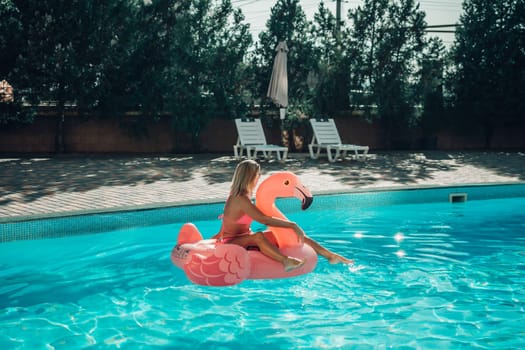 A woman is sitting on a pink flamingo float in a pool. The scene is bright and cheerful, with the woman enjoying her time in the water