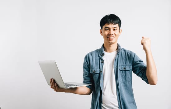 Portrait smiling Asian young man hold a laptop computer studio shot isolated white background, happiness and excited man holding laptop and raising his arm up to celebrate success