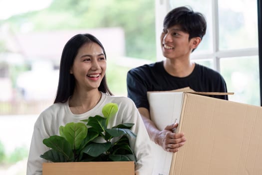 A cheerful couple carrying boxes and plants as they move into their new home, symbolizing a fresh start and new beginnings.