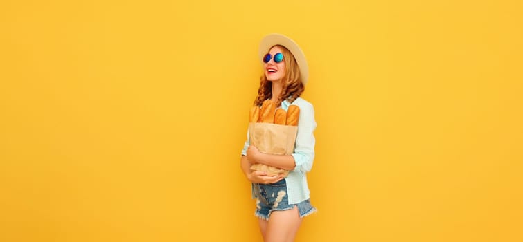 Happy cheerful young woman holding grocery shopping paper bag with fresh long white bread baguette looking away on yellow studio background