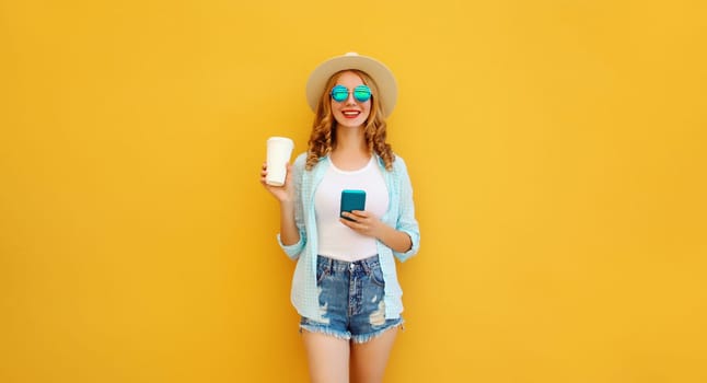 Summer stylish happy young woman with phone, cool girl using smartphone holding cup of coffee dressed tourist straw hat standing on yellow background