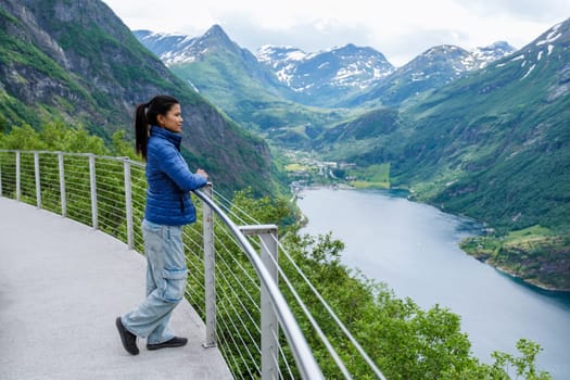 A woman stands on a viewing platform overlooking a fjord in Norway, surrounded by towering green mountains and a clear blue sky. Geiranger fjord