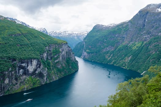 A breathtaking view of a Norwegian fjord surrounded by lush green mountains. The water is a deep blue, reflecting the cloudy sky. Geiranger fjord Norway