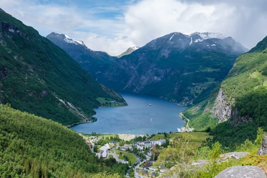 A scenic view of a Norwegian fjord surrounded by towering green mountains. The water is calm and blue, and the sky is clear. Geiranger fjord Norway