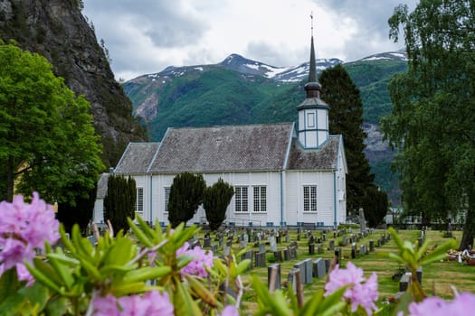 A white church with a steeple stands proudly in a valley in Norway, surrounded by a graveyard and blooming flowers. The majestic mountains provide a breathtaking backdrop. Valldal Norway