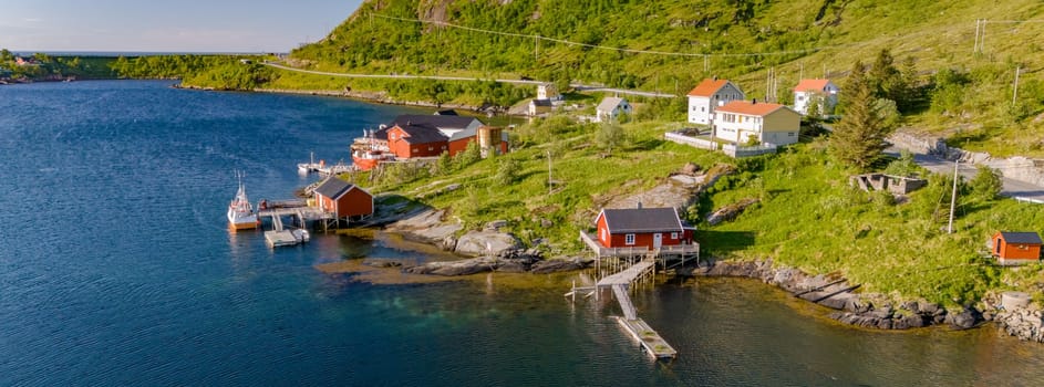 A tranquil scene of red houses nestled on a lush green hillside overlooking a calm blue fjord in Norway. Reine, Lofoten, Norway