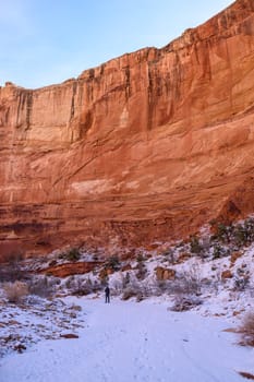 A lone hiker walks through the snowy Grand Wash Trail in Capitol Reef National Park, Utah. The scene showcases striking red rock cliffs under a blue winter sky. Stunning natural beauty - USA