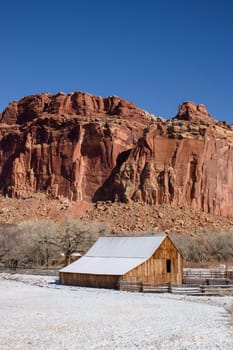 Serene winter scene of a historic wooden barn in Fruita, within Capitol Reef National Park in Utah. The snow-dusted landscape contrasts with the striking red rock cliffs under a bright blue sky - USA