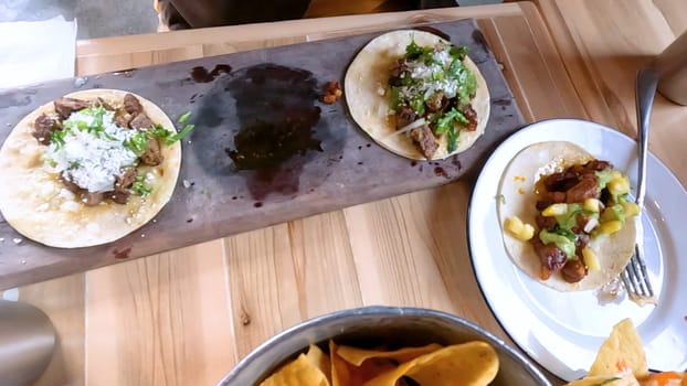 Castle Rock, Colorado, USA-June 12, 2024-Slow motion-Close-up view of a hand picking up a taco from a plate at a restaurant table. The scene features a variety of colorful and appetizing dishes, including nachos topped with cheese and vegetables.