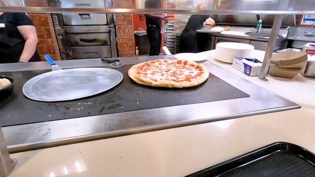 Colorado Springs, Colorado, USA-June 12, 2024-Slow motion-Close-up of a freshly baked pizza slice on a paper plate, placed on a black tray in a pizzeria. The background shows more pizzas being prepared, creating a warm and inviting atmosphere.