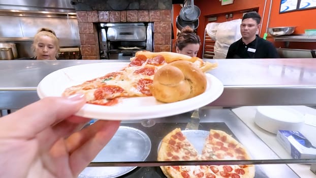 Colorado Springs, Colorado, USA-June 12, 2024-Slow motion-Close-up of a freshly baked pizza slice on a paper plate, placed on a black tray in a pizzeria. The background shows more pizzas being prepared, creating a warm and inviting atmosphere.