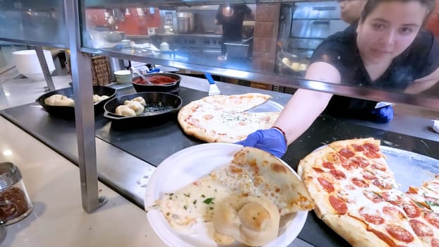 Colorado Springs, Colorado, USA-June 12, 2024-Slow motion-Close-up of a freshly baked pizza slice on a paper plate, placed on a black tray in a pizzeria. The background shows more pizzas being prepared, creating a warm and inviting atmosphere.