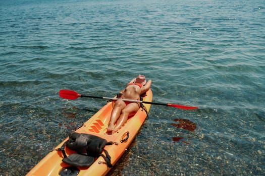 A woman is laying on a kayak in the water. The kayak is orange and has a paddle on it. The woman is wearing a red hat