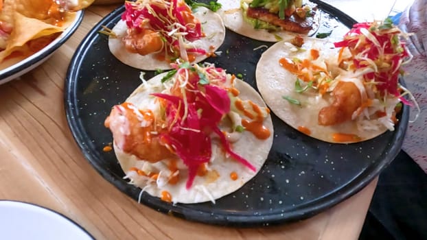 Castle Rock, Colorado, USA-June 12, 2024-Slow motion-Close-up view of a hand picking up a taco from a plate at a restaurant table. The scene features a variety of colorful and appetizing dishes, including nachos topped with cheese and vegetables.