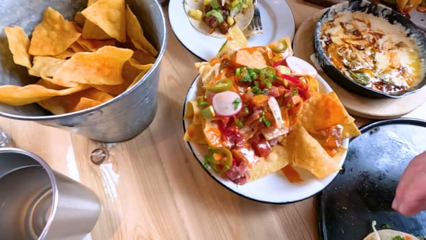 Castle Rock, Colorado, USA-June 12, 2024-Slow motion-Close-up view of a hand picking up a taco from a plate at a restaurant table. The scene features a variety of colorful and appetizing dishes, including nachos topped with cheese and vegetables.