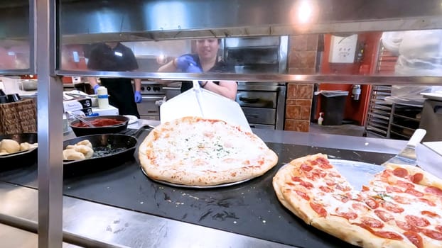 Colorado Springs, Colorado, USA-June 12, 2024-Slow motion-Close-up of a freshly baked pizza slice on a paper plate, placed on a black tray in a pizzeria. The background shows more pizzas being prepared, creating a warm and inviting atmosphere.