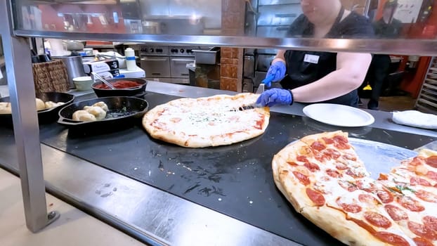 Colorado Springs, Colorado, USA-June 12, 2024-Slow motion-Close-up of a freshly baked pizza slice on a paper plate, placed on a black tray in a pizzeria. The background shows more pizzas being prepared, creating a warm and inviting atmosphere.