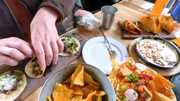 Castle Rock, Colorado, USA-June 12, 2024-Slow motion-Close-up view of a hand picking up a taco from a plate at a restaurant table. The scene features a variety of colorful and appetizing dishes, including nachos topped with cheese and vegetables.