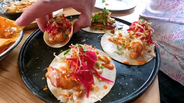 Castle Rock, Colorado, USA-June 12, 2024-Slow motion-Close-up view of a hand picking up a taco from a plate at a restaurant table. The scene features a variety of colorful and appetizing dishes, including nachos topped with cheese and vegetables.