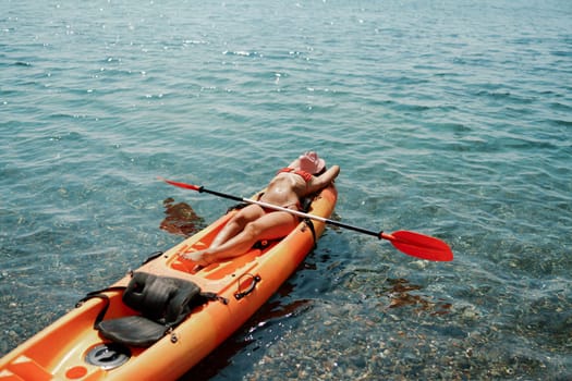 A woman is laying on a kayak in the water. The kayak is orange and has a paddle on it. The woman is wearing a red hat