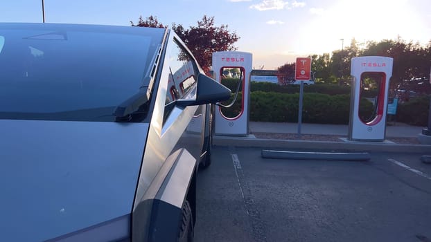 Santa Fe, New Mexico, USA-June 10, 2024-Slow motion-A Tesla Cybertruck is connected to a Tesla Supercharger at an outdoor charging station. The sleek, angular design of the Cybertruck is highlighted by the sunlight, emphasizing its modern and futuristic appearance.