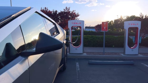 Santa Fe, New Mexico, USA-June 10, 2024-Slow motion-A Tesla Cybertruck is connected to a Tesla Supercharger at an outdoor charging station. The sleek, angular design of the Cybertruck is highlighted by the sunlight, emphasizing its modern and futuristic appearance.