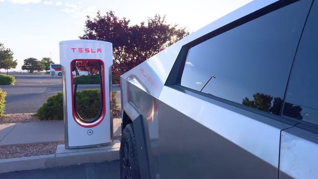 Santa Fe, New Mexico, USA-June 10, 2024-Slow motion-A Tesla Cybertruck is connected to a Tesla Supercharger at an outdoor charging station. The sleek, angular design of the Cybertruck is highlighted by the sunlight, emphasizing its modern and futuristic appearance.