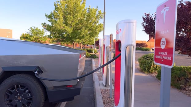 Santa Fe, New Mexico, USA-June 10, 2024-Slow motion-A Tesla Cybertruck is connected to a Tesla Supercharger at an outdoor charging station. The sleek, angular design of the Cybertruck is highlighted by the sunlight, emphasizing its modern and futuristic appearance.